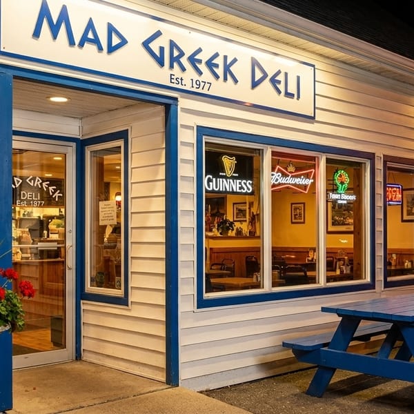 Exterior of Mad Greek Deli at night, with blue picnic tables and neon signs visible through windows.