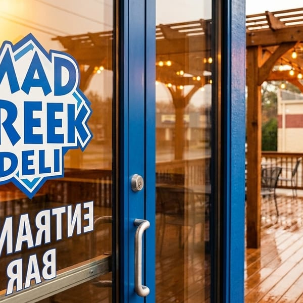 Mad Greek Deli logo on a blue glass door, with an outdoor patio and string lights visible.