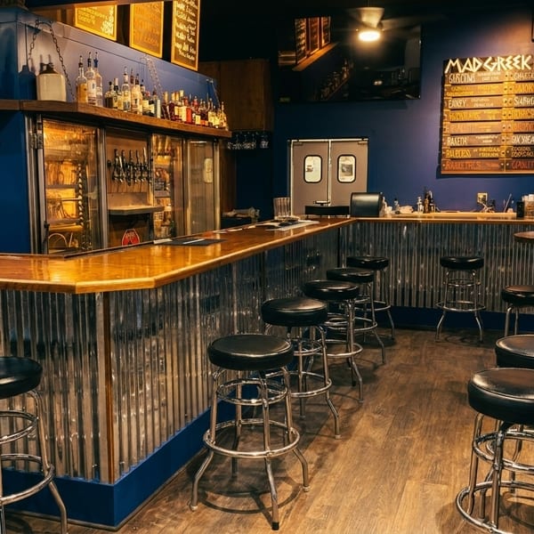 Interior of a bar with a long wooden counter, black stools, and a menu board on a blue wall.