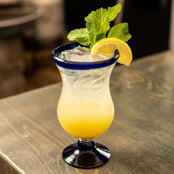Yellow cocktail with ice, lemon slice, and mint in a blue-rimmed glass on a wooden table.