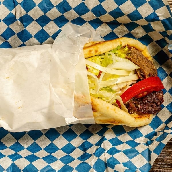 A gyro with meat, lettuce, onions, and red peppers, partially wrapped in white paper on blue checkered paper.