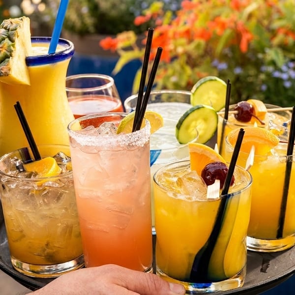 A server carries a tray of colorful cocktails, including a pineapple drink, on an outdoor patio.