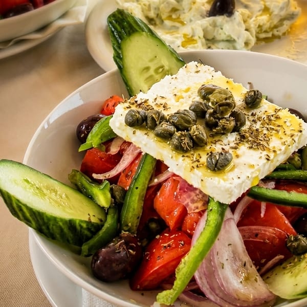 A close-up of a Greek salad with feta cheese, capers, cucumbers, tomatoes, olives, and onions.